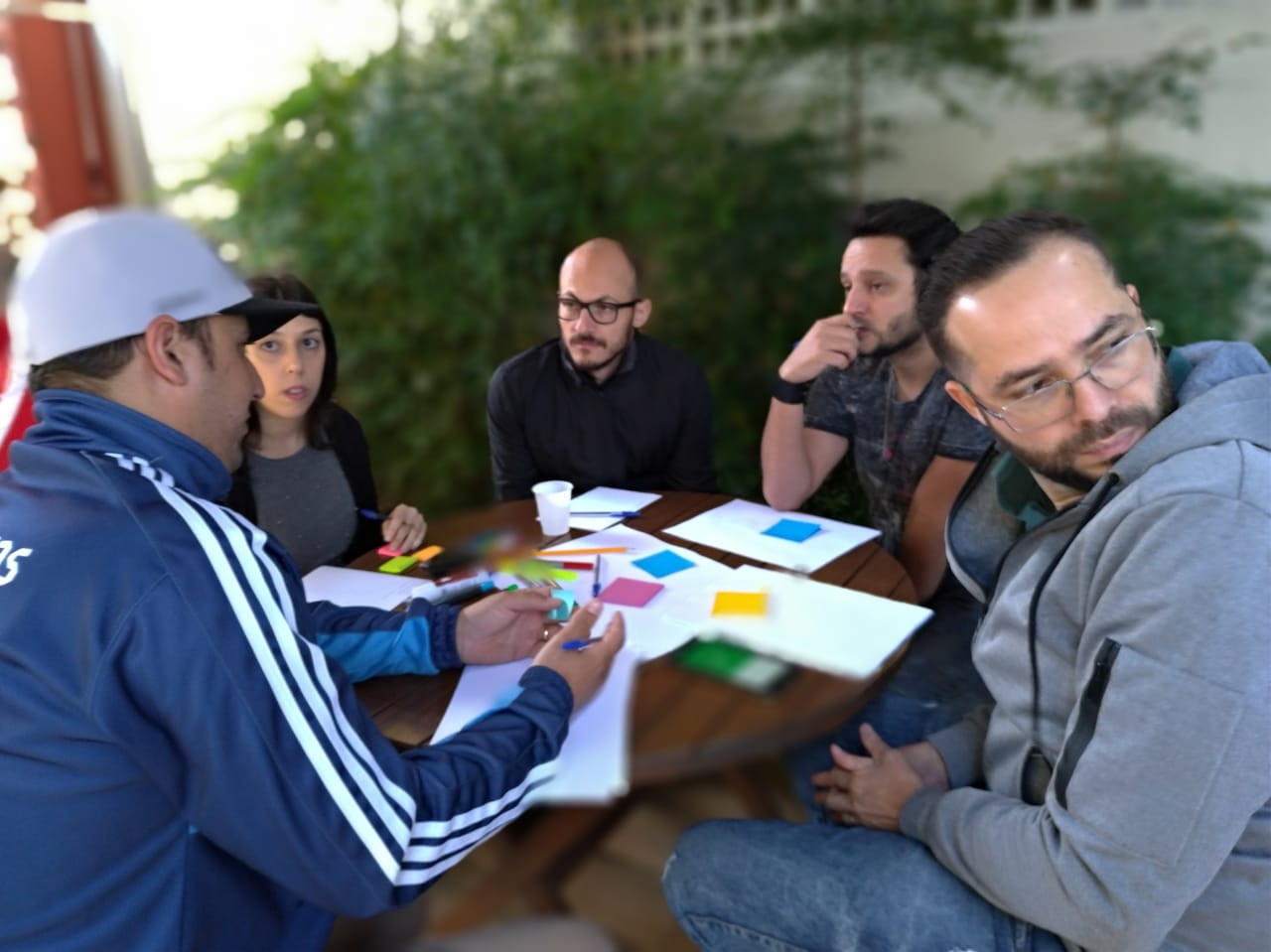 Grupo de cinco pessoas discutindo em torno de uma mesa, com papeis coloridos e anotações; ambiente externo rodeado de vegetação.