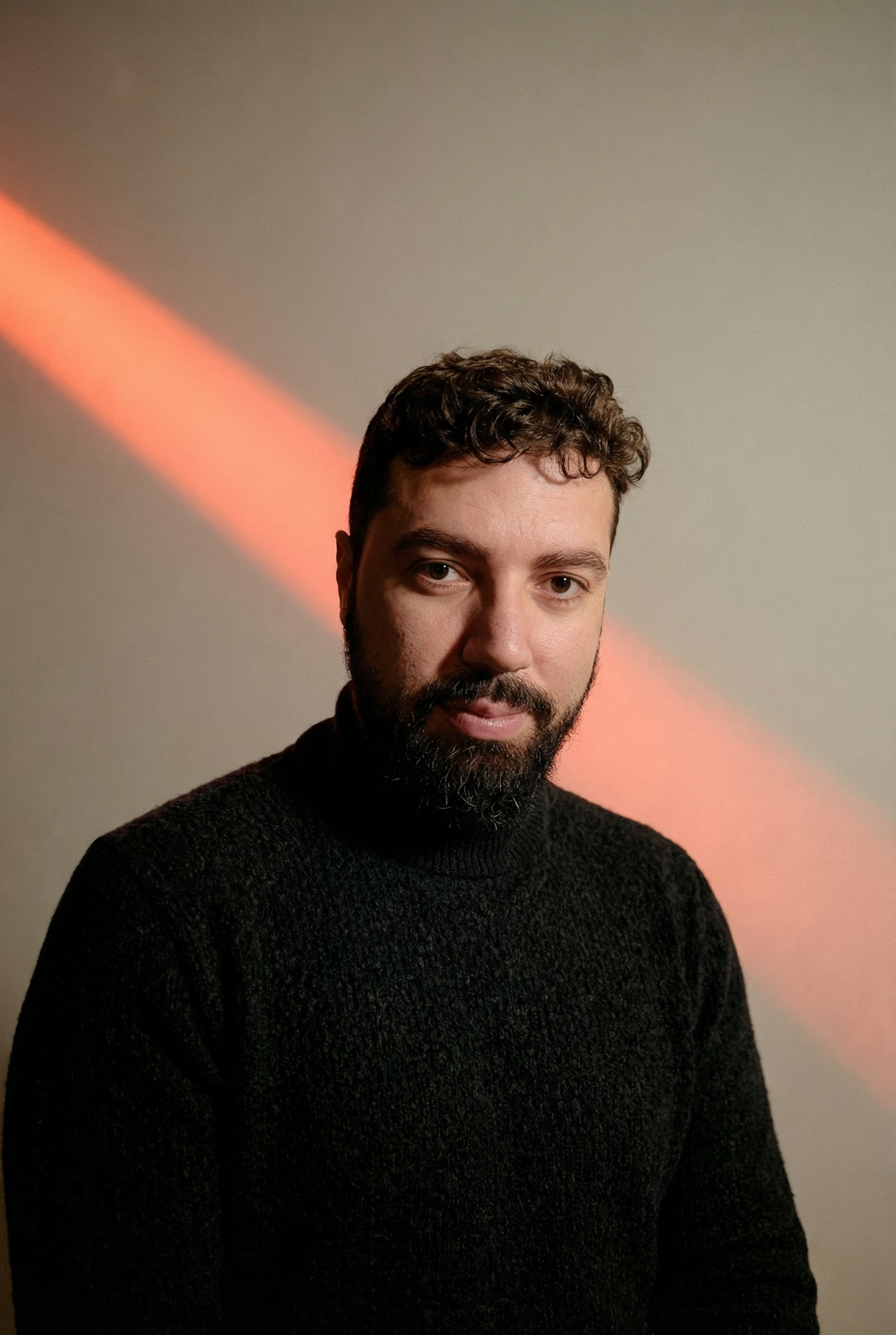Retrato de um homem com barba e cabelo encaracolado, vestindo um suéter escuro, com luz laranja projetada ao fundo.